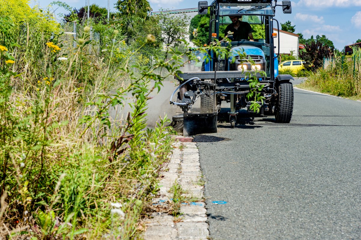 Traktor mit angebauter Wildkrautbürste von matev bei der Arbeit am Straßenrand. Die Bürste entfernt Unkraut entlang einer Bordsteinkante. Im Vordergrund sind hochgewachsene Pflanzen und Wildblumen sichtbar. Der Traktor fährt auf einer asphaltierten Straße, während im Hintergrund Bäume, ein Zaun und ein gelbes Auto zu erkennen sind. Der Himmel ist sonnig und klar.