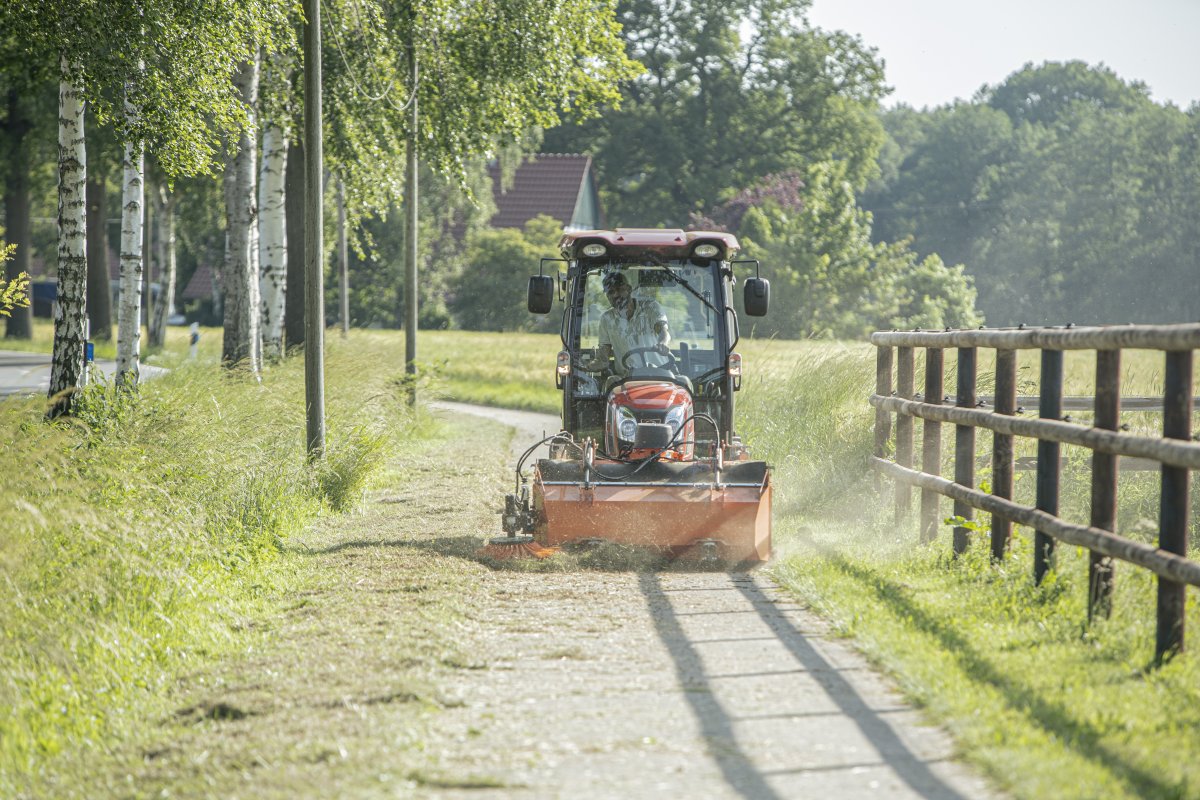 Kioti CX2510 Traktor mit matev Kehrmaschine bei der Reinigung eines schmalen Weges in einer ländlichen Umgebung. Baumreihe und Holzzaun im Hintergrund, ideale Lösung für die professionelle Wegepflege.