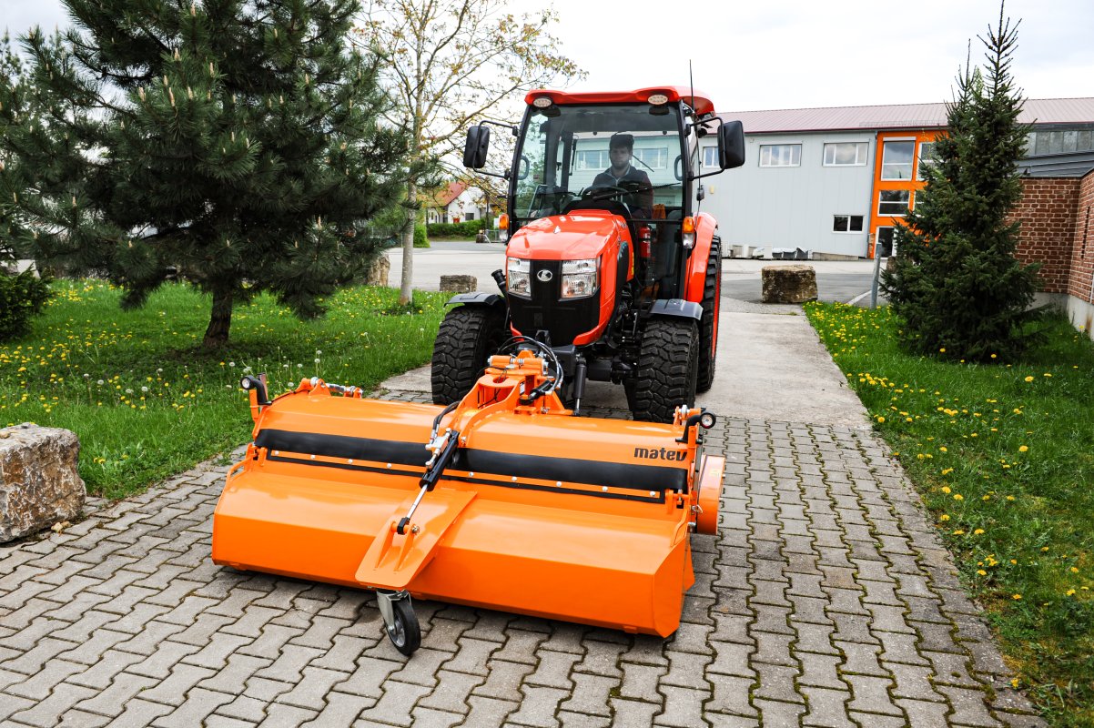 Kubota L2-622 Traktor mit angehängter matev Kehrmaschine in leuchtendem Orange, ideal für professionelle Reinigung von gepflasterten Wegen und Einfahrten. Im Hintergrund moderne Gebäude und Grünflächen mit Bäumen.