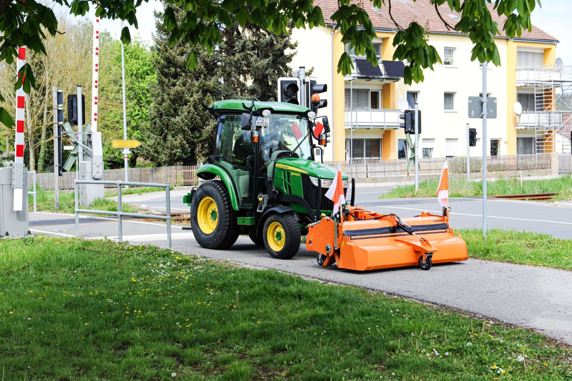 John Deere 3R Traktor mit matev Kehrmaschine in Orange bei der Reinigung eines Gehwegs neben einer Bahnschranke und Wohnhäusern. Perfekte Lösung für die Pflege von Gehwegen und urbanen Flächen.