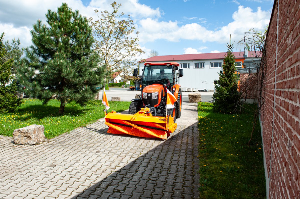 Kubota L2-622 Traktor mit matev Kehrmaschine in Orange, bei der Reinigung eines gepflasterten Weges neben einem Backsteingebäude. Perfekt für die professionelle Reinigung von Wegen und Zufahrten in städtischer oder ländlicher Umgebung.