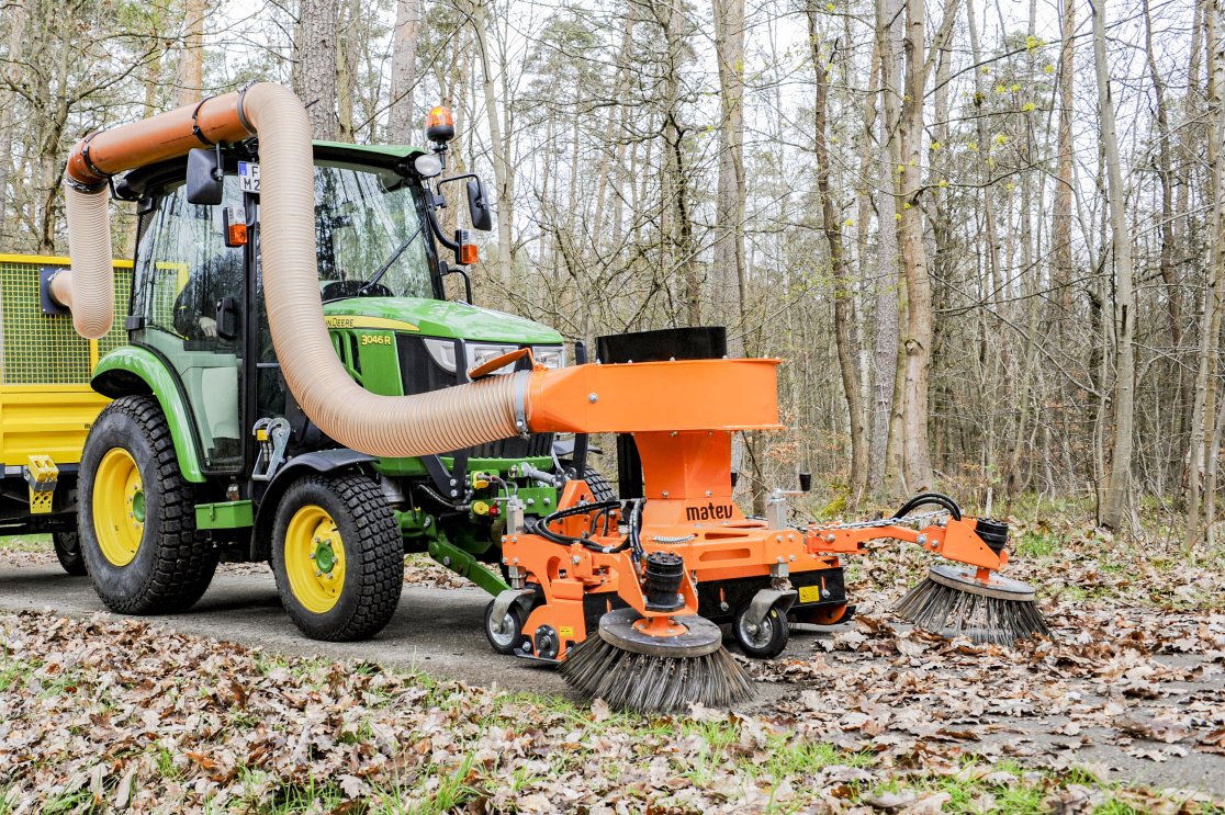 Grüner John Deere Traktor Modell 3046R, ausgestattet mit dem orangefarbenen matev MRM-120 AS Saugkehrsystem und einer Kehrbürste, bei der Reinigung eines Laubbedeckten Weges in einem Waldgebiet.