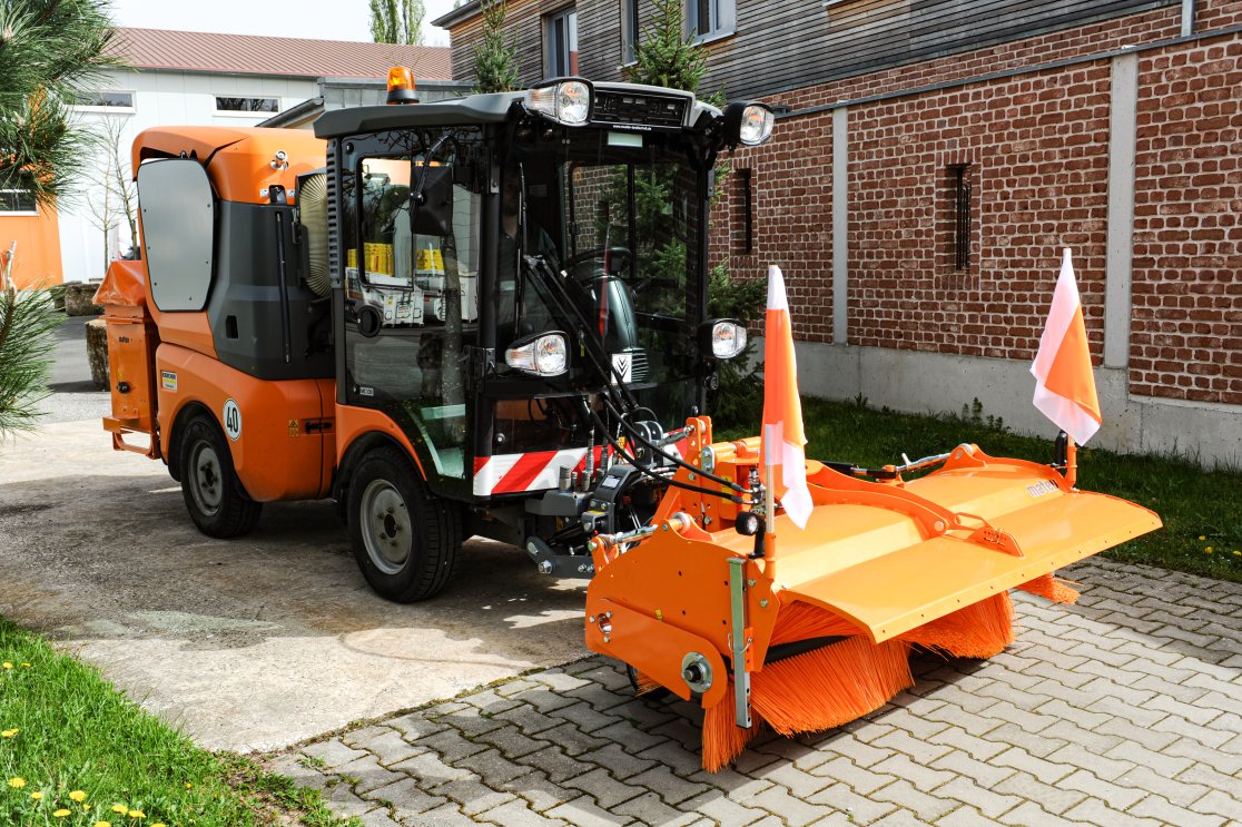 Eine orangefarbene Kommunalmaschine Kärcher MC 130, ausgestattet mit einer matev SWE 14-57 Kehrmaschine und einem Kastenstreuer. Die Maschine steht auf einem gepflasterten Hof vor einer Backsteinwand, im Hintergrund sind grüne Bäume und ein Gebäude zu sehen.