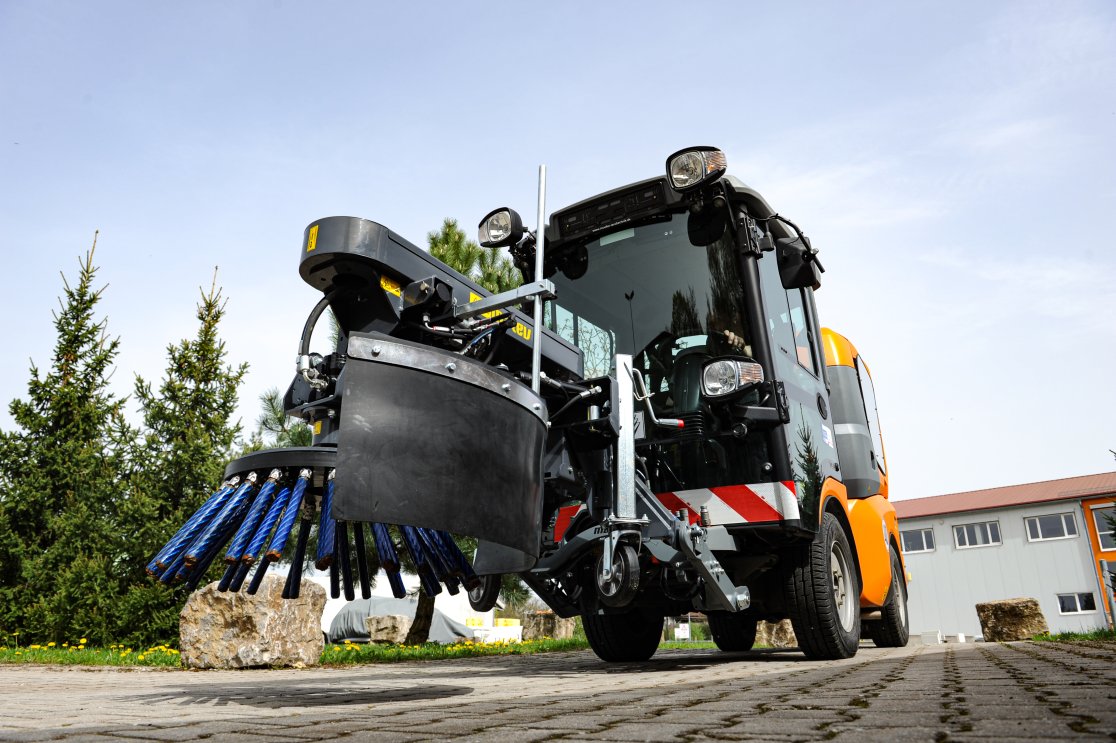 Wildkrautbürste im Einsatz. Im Vordergrund ist die blaue Bürste zu sehen, die sich auf einer gepflasterten Fläche befindet. Im Hintergrund stehen Bäume, ein Gebäude und ein klarer Himmel.