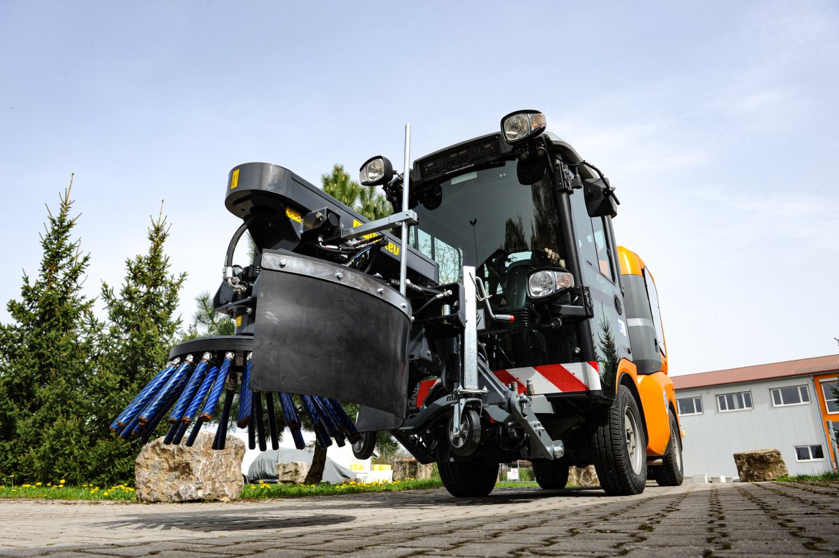Wildkrautbürste im Einsatz. Im Vordergrund ist die blaue Bürste zu sehen, die sich auf einer gepflasterten Fläche befindet. Im Hintergrund stehen Bäume, ein Gebäude und ein klarer Himmel.