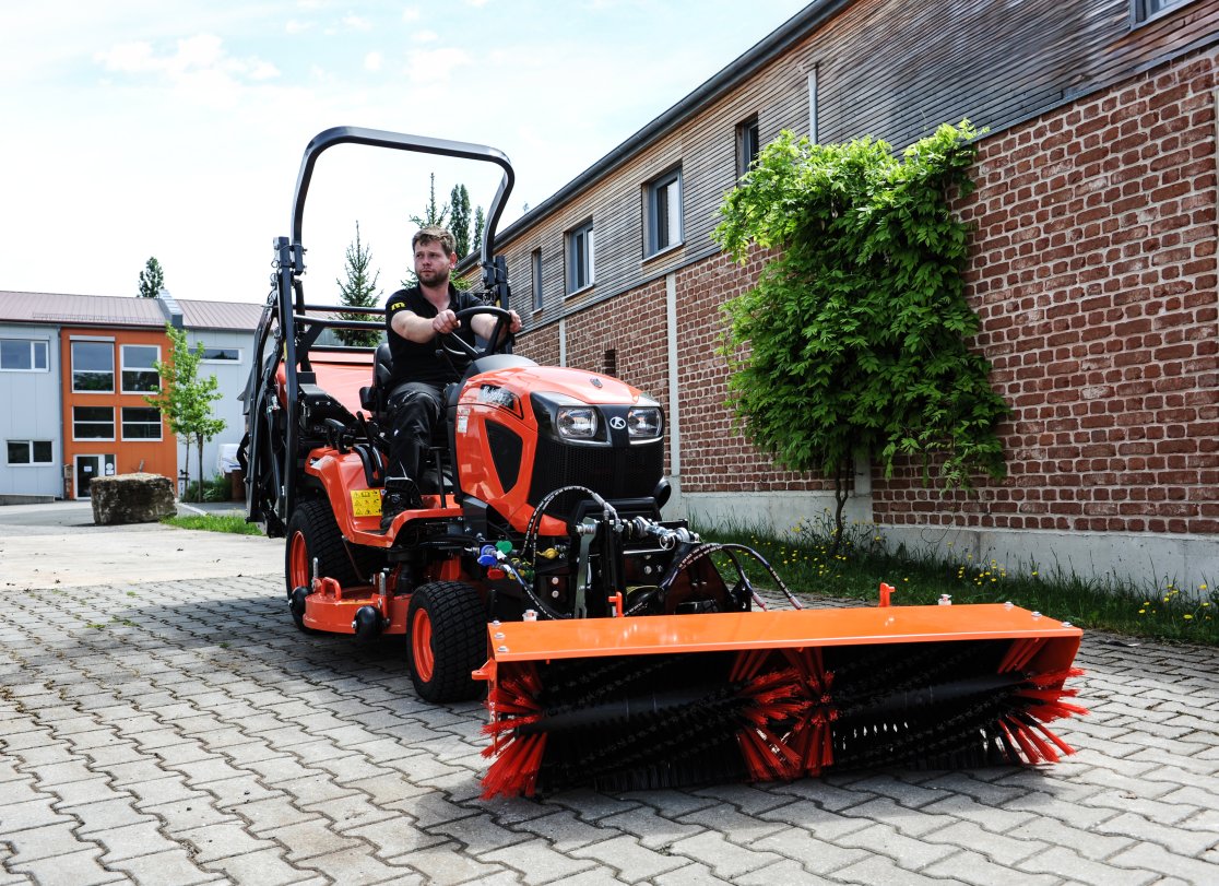 Ein Kubota G261 Traktor mit angebauter matev Kehrmaschine SWE KU 140. Ein Mann sitzt auf dem Fahrzeug und steuert es. Die Kehrmaschine mit roten Bürsten ist auf gepflastertem Boden vor einer roten Ziegelmauer mit grünen Pflanzen zu sehen. Im Hintergrund befinden sich Gebäude und ein kleiner Baum.