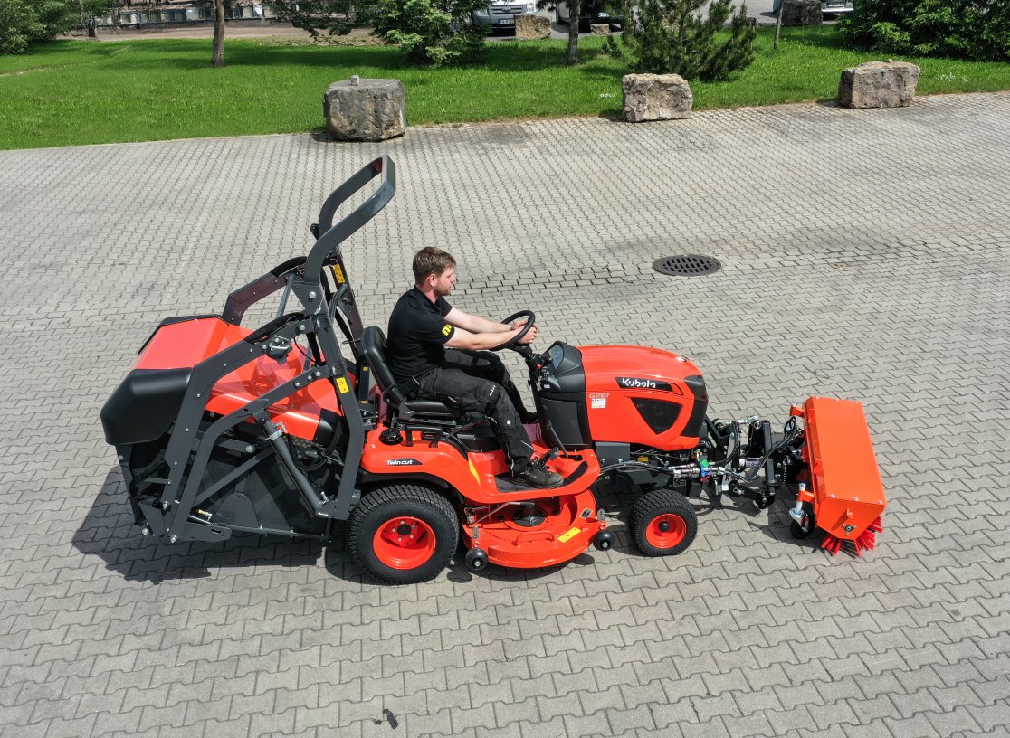 Ein Kubota G261 mit angebauter matev Kehrmaschine SWE KU 140. Ein Bediener sitzt auf dem roten Fahrzeug, das auf einem gepflasterten Hofgelände geparkt ist. Die Kehrmaschine ist orangefarben und zeigt ihre rotierenden Bürsten. Im Hintergrund ist eine grüne Rasenfläche mit Bäumen und Steinen sichtbar.