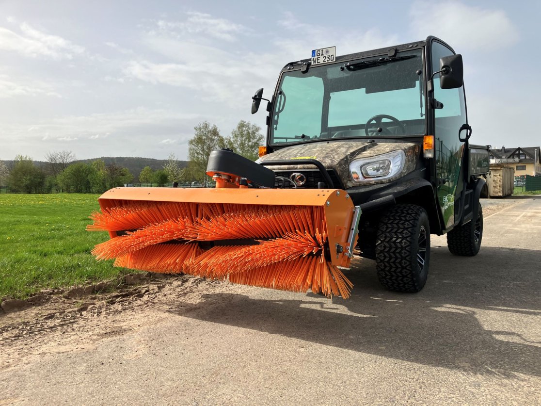 Ein Kubota RTV in Camouflage-Optik mit einer angebauten matev Kehrmaschine SWE 16/45. Die orangefarbenen Bürsten der Kehrmaschine heben sich kontrastreich vom grünen Gras im Hintergrund ab. Das Fahrzeug steht auf einem befestigten Weg mit leicht bewölktem Himmel und umgebender Natur.