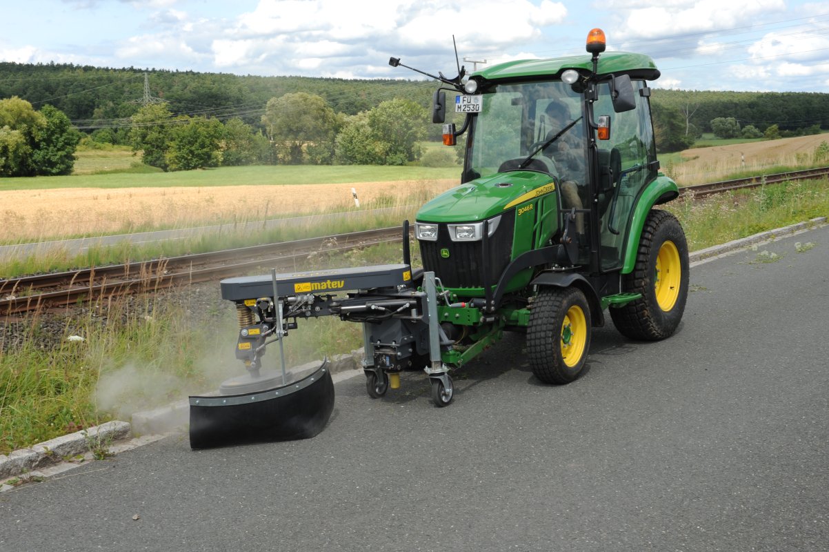 Traktor John Deere 3046R mit angebauter Wildkrautbürste von matev bei der Arbeit am Straßenrand. Die Bürste entfernt Unkraut, während im Hintergrund Felder, Bäume und ein Bahngleis zu sehen sind. Der Himmel ist teils bewölkt.