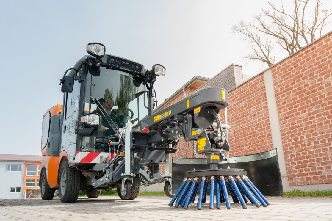 Nahaufnahme einer angebauten Wildkrautbürste von matev auf einer gepflasterten Fläche. Die blauen Bürstenborsten und die robuste Konstruktion sind deutlich sichtbar. Im Hintergrund eine Ziegelmauer, ein modernes Gebäude und ein klarer Himmel. Der Fahrer ist durch die Kabine erkennbar.