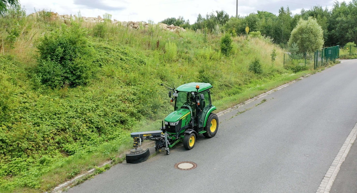 Vogelperspektive auf einen John Deere 3046R Traktor mit angebauter Wildkrautbürste von matev, der am Straßenrand Unkraut entfernt. Im Hintergrund ist eine bewachsene Böschung mit Sträuchern und Bäumen zu sehen. Der Himmel ist leicht bewölkt.