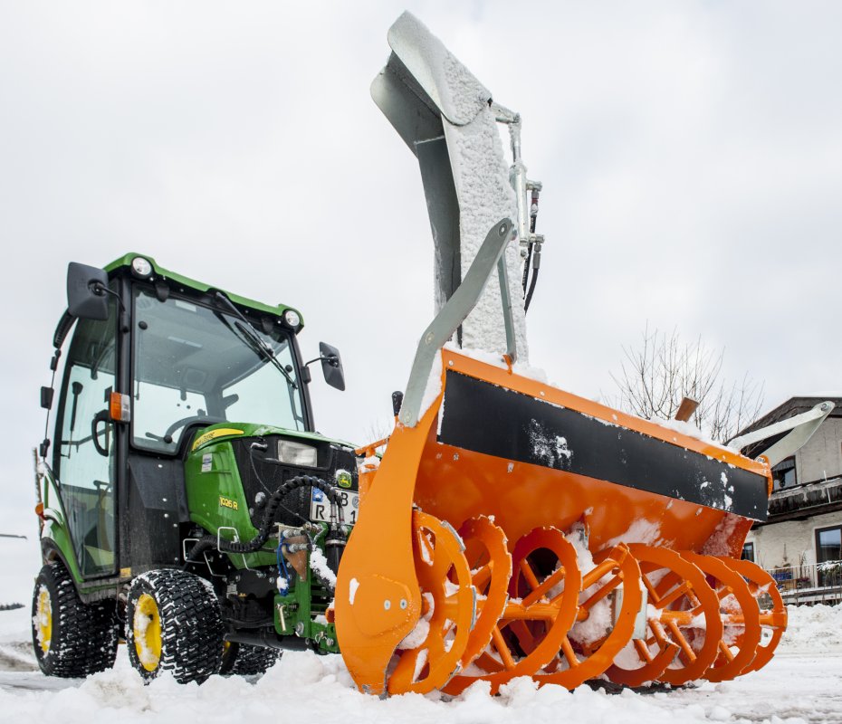 Nahaufnahme eines John Deere Traktors 1026R mit angebauter orangefarbener matev-Schneefräse. Die Schneefräse ist in Aktion und zeigt ihre robuste Bauweise mit den Frässchnecken. Der Traktor steht auf einer schneebedeckten Fläche, die Reifen sind mit Winterprofil versehen. Im Hintergrund sind kahle Bäume und ein Gebäude unter bewölktem Himmel zu sehen.