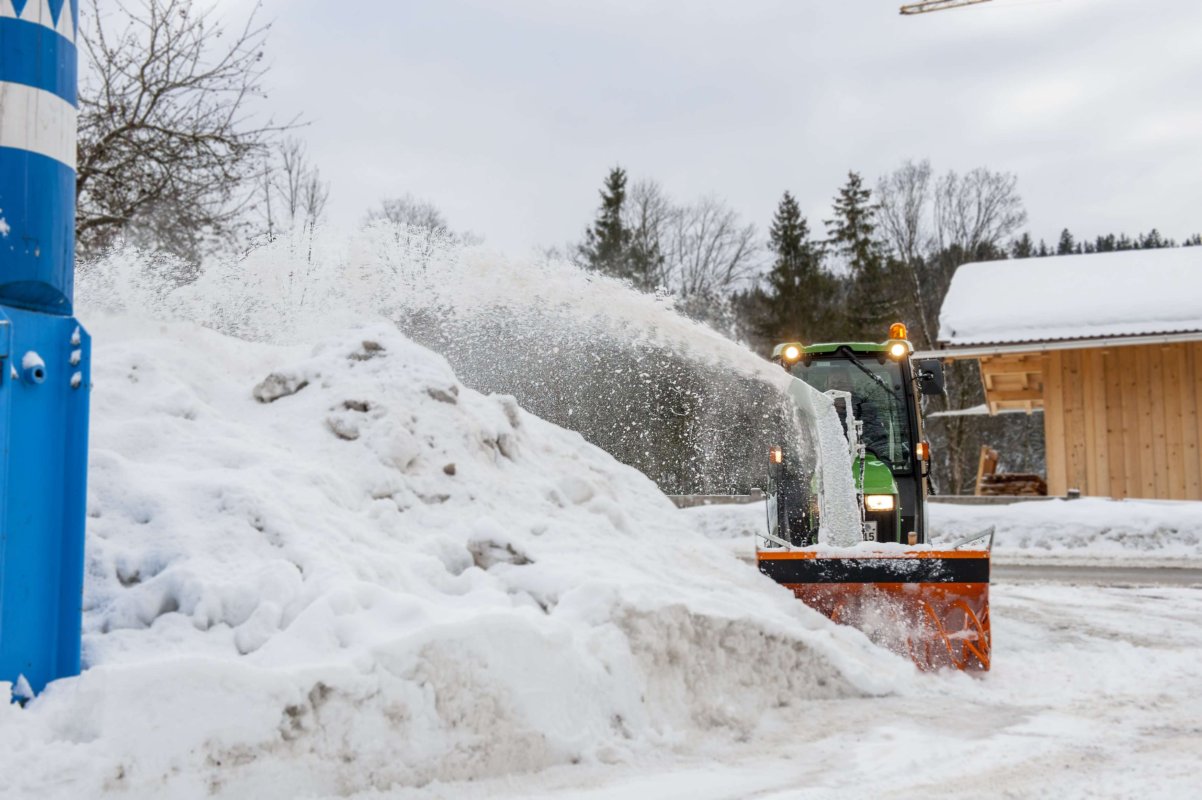 John Deere Traktor mit angebauter matev-Schneefräse im Einsatz. Die Schneefräse wirft Schnee seitlich in einem hohen Bogen. Im Vordergrund liegt eine große Schneeansammlung, und im Hintergrund sind ein Maibaum, Bäume und ein Holzgebäude zu sehen. Die Szene zeigt eine verschneite Winterlandschaft unter bewölktem Himmel.