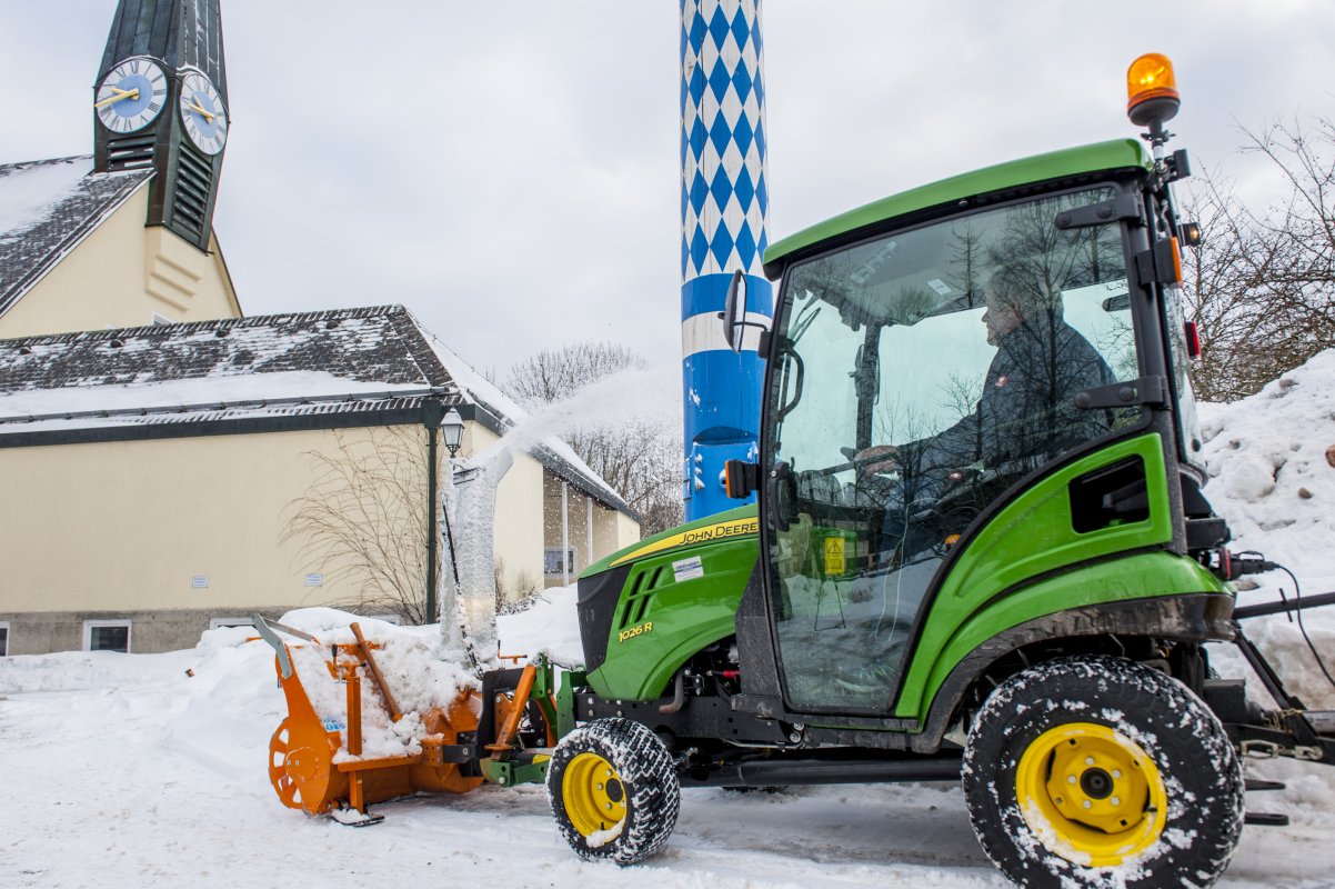 John Deere Traktor 1026R mit angebauter orangefarbener matev-Schneefräse im Einsatz. Die Schneefräse wirft Schnee seitlich aus. Im Hintergrund sind eine Kirche mit Uhrturm, ein traditioneller Maibaum und schneebedeckte Flächen sichtbar. Der Fahrer sitzt in der beheizten Kabine, und die Umgebung zeigt eine winterliche Dorfkulisse.