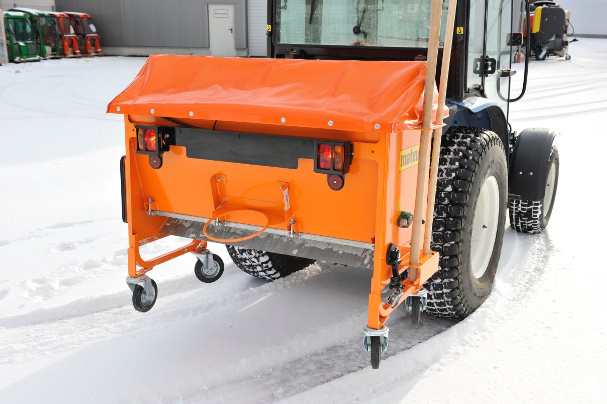 Rückansicht eines Traktors mit angebautem orangefarbenem matev-Kastenstreuer BX. Der Streuer hat eine Abdeckplane und steht auf einer verschneiten Fläche. Im Hintergrund sind weitere Arbeitsmaschinen und ein Gebäude erkennbar. Die Traktorreifen haben ein wintertaugliches Profil, und die Umgebung ist von Schnee bedeckt.