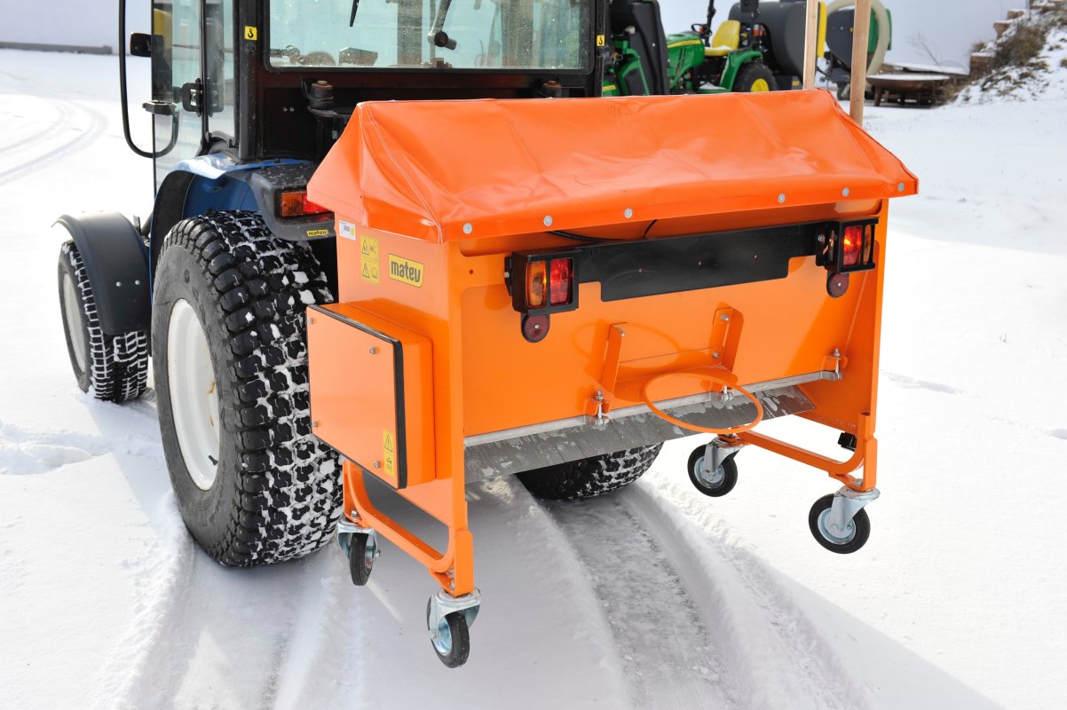 Rückansicht eines Traktors mit angebautem orangefarbenem matev-Kastenstreuer BX auf einer verschneiten Fläche. Der Streuer ist mit einer Abdeckplane und Rückleuchten ausgestattet. Im Hintergrund sind weitere landwirtschaftliche Fahrzeuge zu sehen. Die Umgebung ist winterlich, mit Schnee bedeckten Flächen und Traktorreifen mit Winterprofil.
