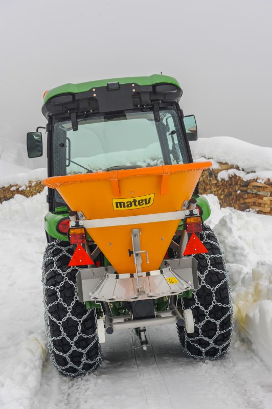 Rückansicht eines John Deere Traktors mit angebautem orangefarbenem matev-Schleuderstreuer DS. Die Traktorreifen sind mit Schneeketten ausgestattet, und der Traktor steht auf einem schneebedeckten Weg. Im Hintergrund liegen Holzstapel, und die Umgebung ist von dichtem Schnee und Nebel geprägt.