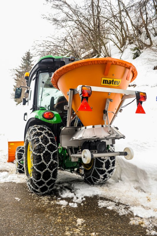 Rückansicht eines John Deere Traktors mit angebautem orangefarbenem matev-Schleuderstreuer DS auf einem schneebedeckten Weg. Die Traktorreifen sind mit Schneeketten ausgestattet, und im Hintergrund befinden sich verschneite Hänge und Bäume. Das orangefarbene Schneeräumschild ist an der Vorderseite des Traktors sichtbar. Die Szene zeigt eine winterliche Umgebung.