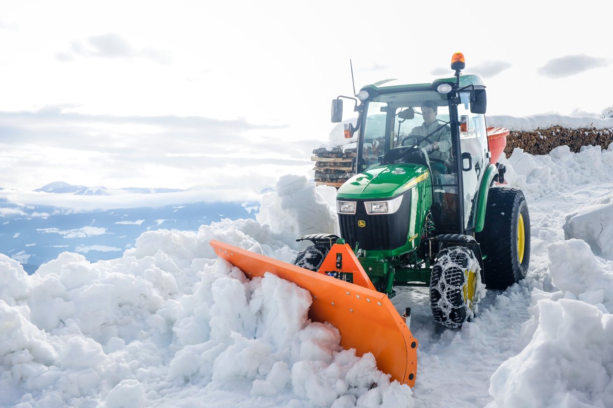 Ein John Deere 4066R Traktor mit einem orangefarbenen SRM-SF Schneeräumschild von matev schiebt Schnee auf einer verschneiten Fläche. Der Traktor ist mit Schneeketten ausgestattet, im Hintergrund eine Berglandschaft und gestapeltes Brennholz.