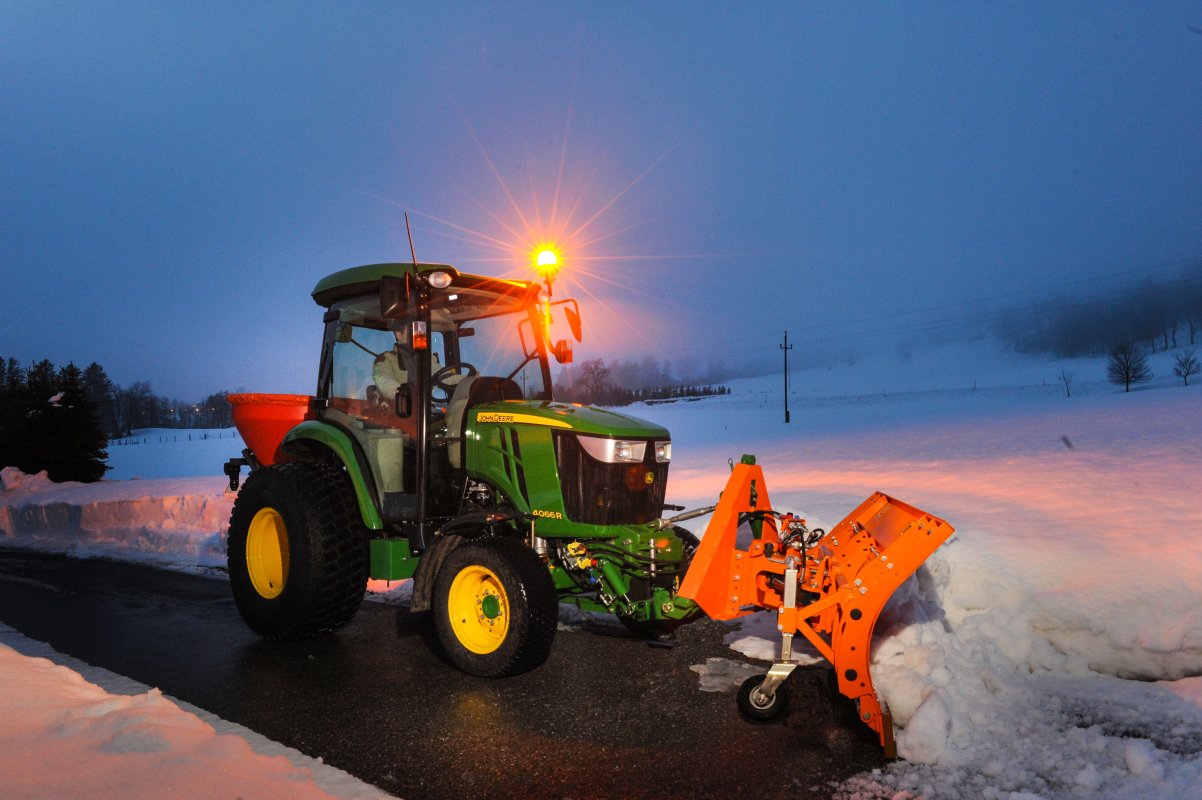 "Ein John Deere 4066R Traktor mit geschlossenem Führerhaus und eingeschaltetem orangefarbenem Warnlicht bei Dämmerung. Ausgestattet mit einem Schneeräumschild und einem Streugerät, räumt der Traktor Schnee auf einer asphaltierten Straße in einer verschneiten Landschaft.