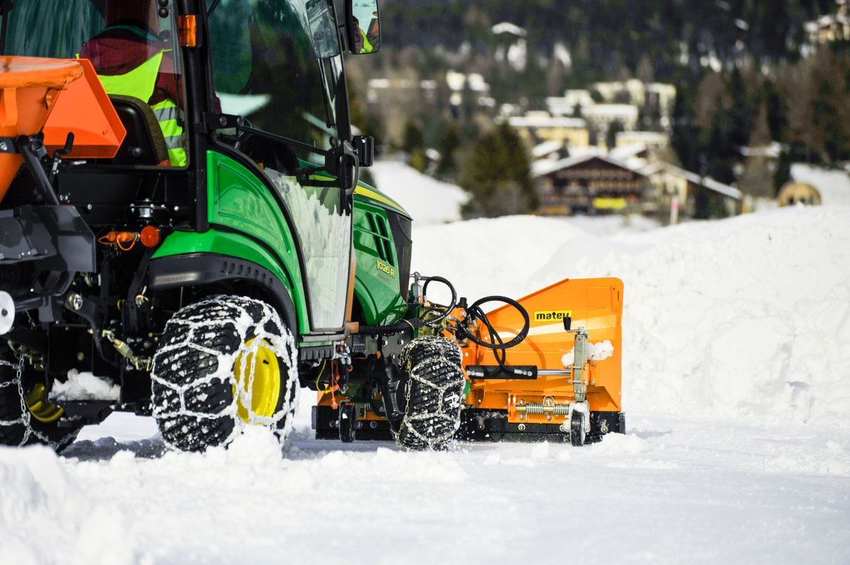 Ein John Deere 1026R Kompakttraktor mit Schneeketten und einem orangefarbenen matev SRM-VP Schneepflug. Der Traktor ist im Einsatz auf einer schneebedeckten Straße, im Hintergrund sind Gebäude und winterliche Landschaft zu sehen.