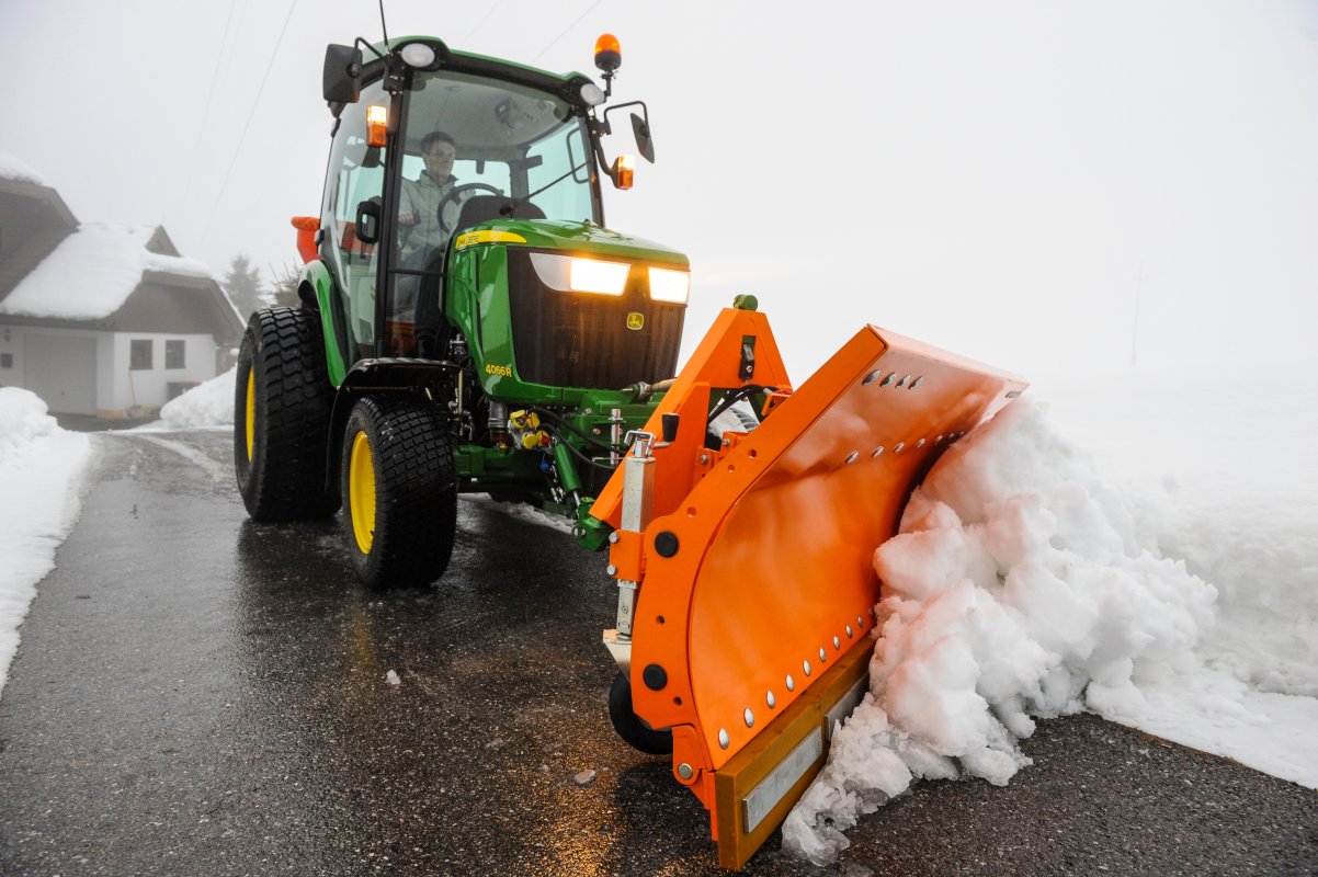 Ein John Deere 4066R Traktor mit eingeschaltetem Licht und einem orangefarbenen SRM-SF Schneeräumschild von matev räumt Schnee von einer asphaltierten Straße. Im Hintergrund sind schneebedeckte Gebäude und neblige Winterstimmung zu sehen.