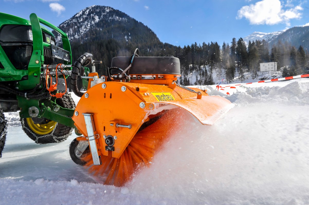 Eine matev-Kehrmaschine, montiert an einem grünen John Deere Traktor, fegt Schnee auf einer verschneiten Fläche. Die orangefarbenen Bürsten wirbeln den Schnee kraftvoll auf. Im Hintergrund erstreckt sich eine Winterlandschaft mit schneebedeckten Bergen und Wäldern unter einem strahlend blauen Himmel. Die Szene zeigt den professionellen Einsatz der Kehrmaschine im Winterdienst.