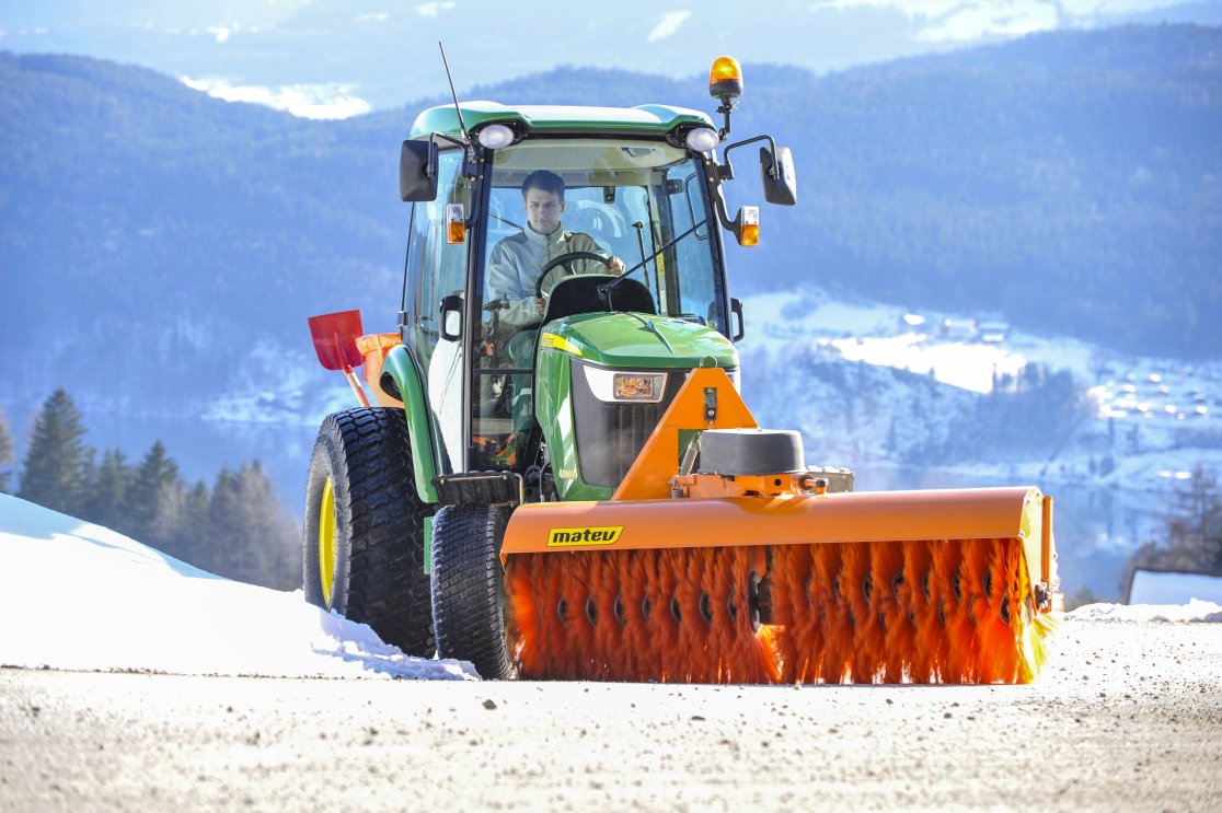 John Deere Traktor 4066R mit angebauter orangefarbener matev-Kehrmaschine SWE-60 im Einsatz auf einer schneebedeckten Straße. Der Fahrer arbeitet konzentriert in der beheizten Kabine. Im Hintergrund erstreckt sich eine winterliche Berglandschaft mit Bäumen und verschneiten Häusern. Die Szene zeigt einen klaren, sonnigen Wintertag.