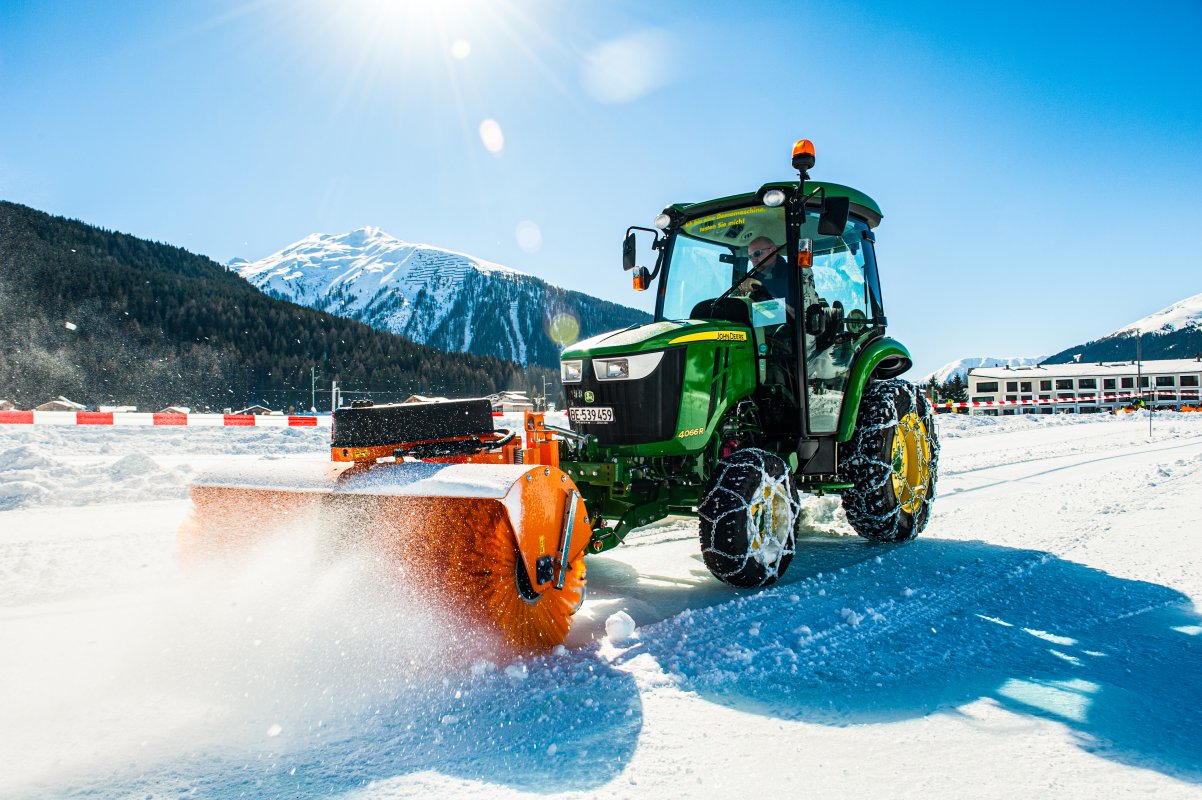 John Deere Traktor 4066R mit angebauter matev-Kehrmaschine SWE-60 im Einsatz auf einer schneebedeckten Fläche unter strahlendem Sonnenschein. Die Schneebürste wirbelt Schnee auf, während der Traktor mit Schneeketten ausgerüstet ist. Im Hintergrund sind schneebedeckte Berge, ein Wald und ein modernes Gebäude zu sehen. Die Szene zeigt einen klaren Wintertag in alpiner Umgebung.