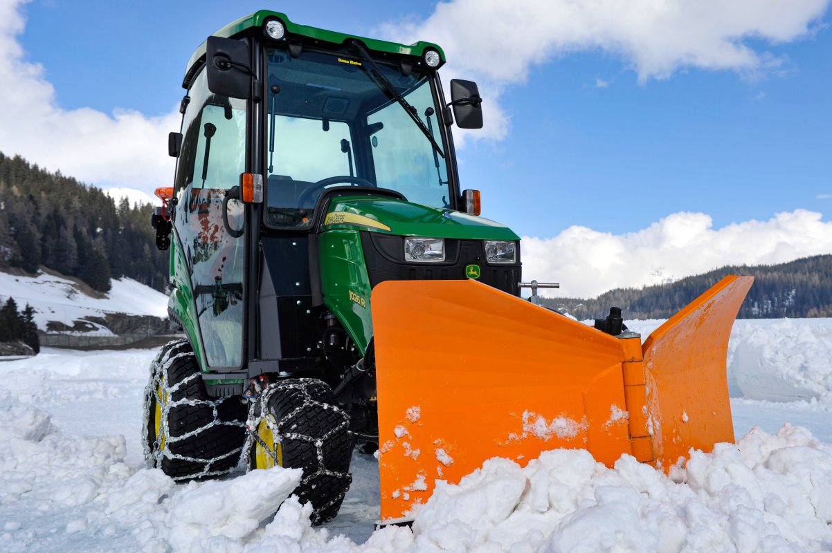 Ein John Deere 1026R Kompakttraktor mit einer geschlossenen Kabine und Schneeketten, ausgestattet mit einem orangefarbenen Schneepflug, räumt Schnee in einer winterlichen Berglandschaft. Der klare Himmel und verschneite Hügel bilden die Kulisse.