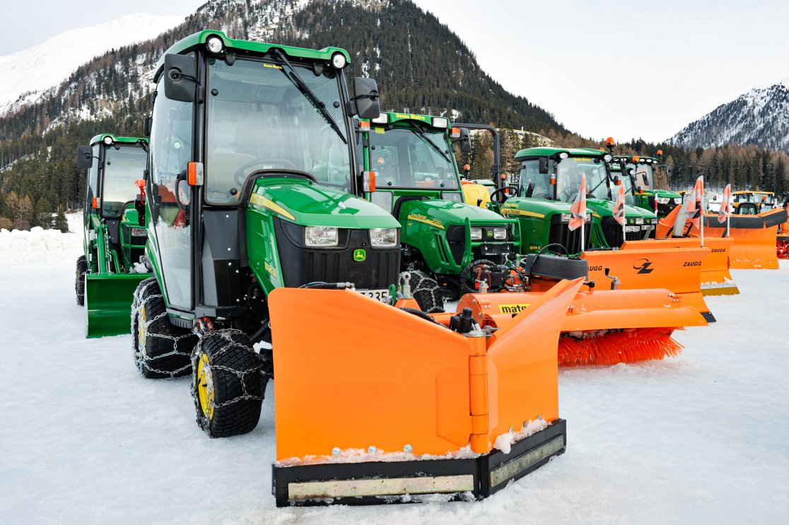 Eine Reihe von John Deere Traktoren mit geschlossenen Kabinen, Schneeketten und verschiedenen Winteranbaugeräten wie matev Schneepflügen und Kehrmaschinen, präsentiert auf einer schneebedeckten Fläche in einer malerischen Berglandschaft.