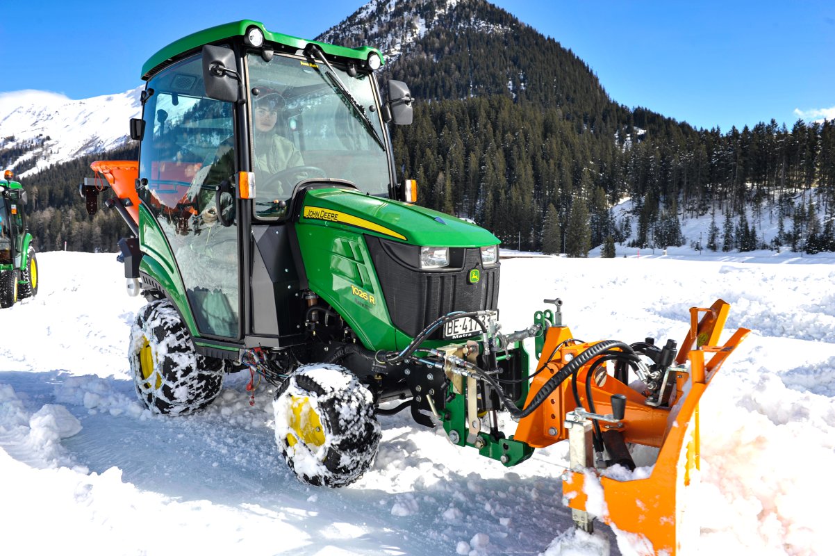 Ein grüner John Deere 1026R Kompakttraktor mit einem orangefarbenen SRM-VP Schneepflug von matev im Einsatz auf verschneitem Gelände vor einer Bergkulisse. Der Traktor ist mit Winterreifen und einem Streugerät ausgestattet und sorgt für eine professionelle Schneeräumung.