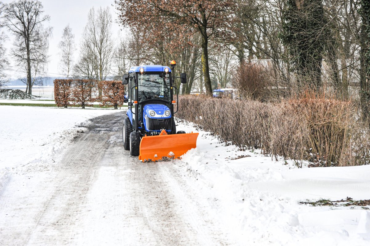 Ein blauer New Holland Traktor mit einem orangefarbenen SRM-FB Schneeräumschild von matev räumt Schnee auf einem schmalen Weg. Der Weg ist von kahlen Bäumen und Hecken gesäumt, die winterliche Landschaft im Hintergrund ergänzt das Bild.