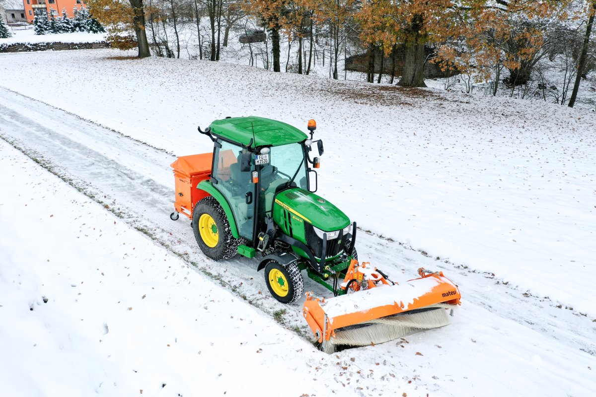 ohn Deere Traktor 3046R mit angebauter matev-Kehrmaschine SWE-57 und Kastenstreuer BX-100 im Wintereinsatz. Der Traktor reinigt einen schneebedeckten Weg, während der Kastenstreuer Salz oder Streugut bereitstellt. Die Umgebung zeigt eine verschneite Wiese mit Bäumen und Laub sowie ein Gebäude im Hintergrund. Die Szene vermittelt einen typischen Winterarbeitsvorgang.
