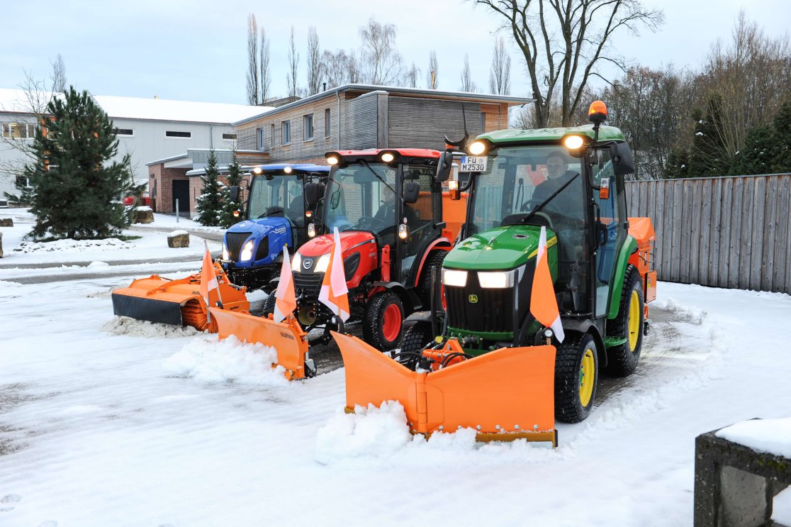 Drei kompakte Traktoren verschiedener Marken (blau, rot und grün) ausgestattet mit orangefarbenen matev Schneepflügen und Kehrsystemen im Winterdienst-Einsatz auf schneebedecktem Gelände, vor einem modernen Firmengebäude mit Bäumen im Hintergrund.