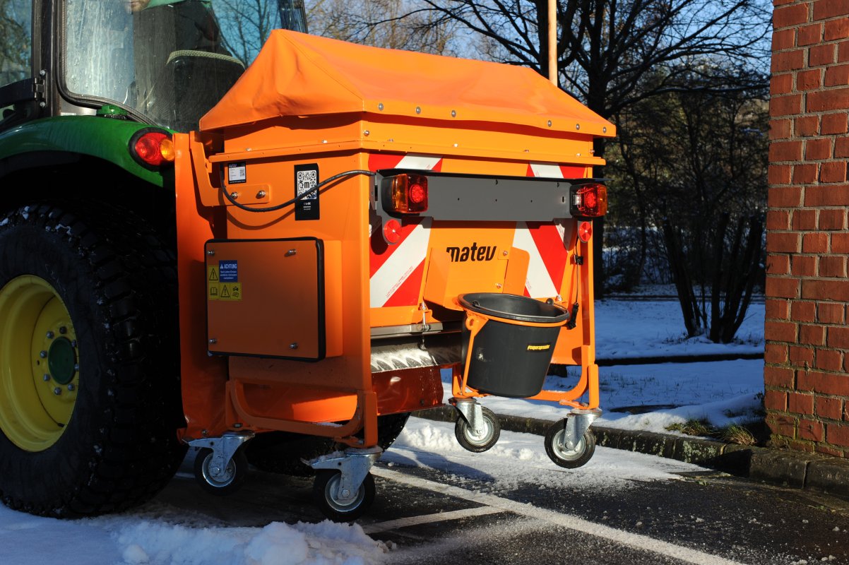 Rückansicht eines John Deere Traktors mit angebautem matev-Streuer BX-100. Der orangefarbene Streuer mit Abdeckplane und Warnmarkierungen ist auf einer winterlichen Straße abgestellt. Ein schwarzer Eimer ist an der Rückseite befestigt. Im Hintergrund sind schneebedeckte Flächen, Bäume und eine Ziegelmauer sichtbar.