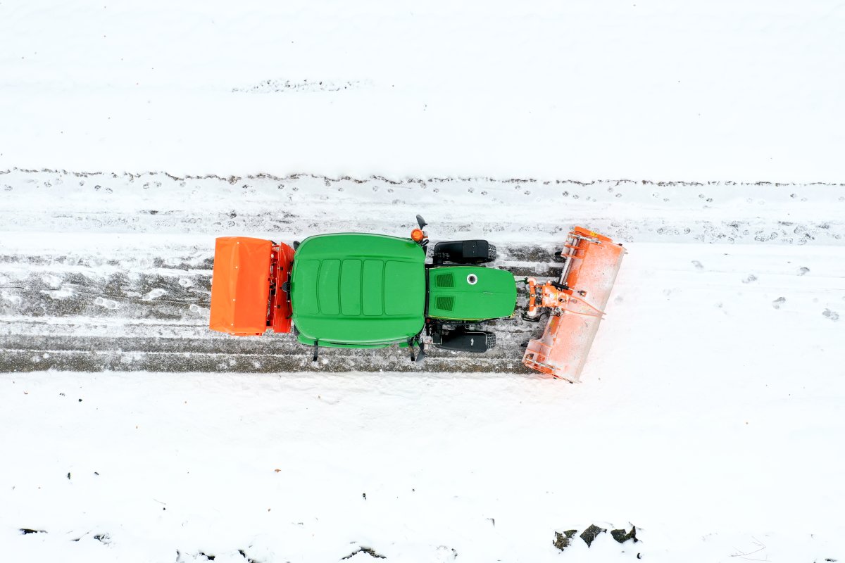 Vogelperspektive eines John Deere Traktors 3046R mit angebauter orangefarbener matev-Kehrmaschine SWE-57 und Kastenstreuer BX-100. Der Traktor arbeitet auf einem schneebedeckten Weg, der teilweise geräumt ist. Die Umgebung zeigt eine weiße Winterlandschaft mit Spuren im Schnee.