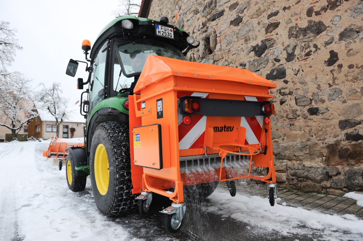 Rückansicht eines John Deere Traktors mit angebautem orangefarbenem matev-Kastenstreuer BX-100 beim Streuen von Salz auf einer verschneiten Straße. Der Streuer ist mit Warnmarkierungen und Abdeckplane ausgestattet. Im Hintergrund sind eine historische Steinmauer, Gebäude und schneebedeckte Bäume zu sehen. Die Szene zeigt einen winterlichen Einsatz.