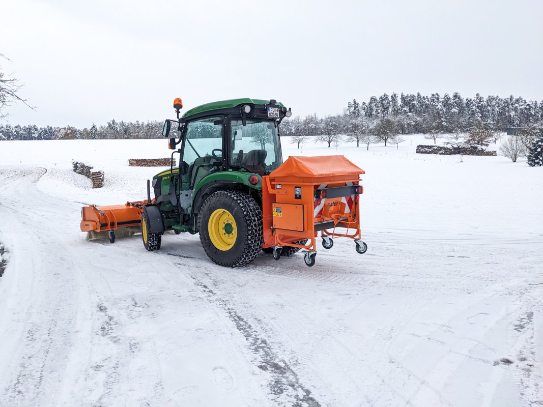 John Deere Traktor mit angebautem matev-Kastenstreuer BX und Kehrbürste im Einsatz auf einem schneebedeckten Weg. Der Traktor ist mit Winterreifen ausgestattet, und die Umgebung zeigt eine verschneite Landschaft mit Bäumen und Holzstapeln im Hintergrund. Der Himmel ist bewölkt, und die Szene vermittelt einen winterlichen Arbeitseinsatz.