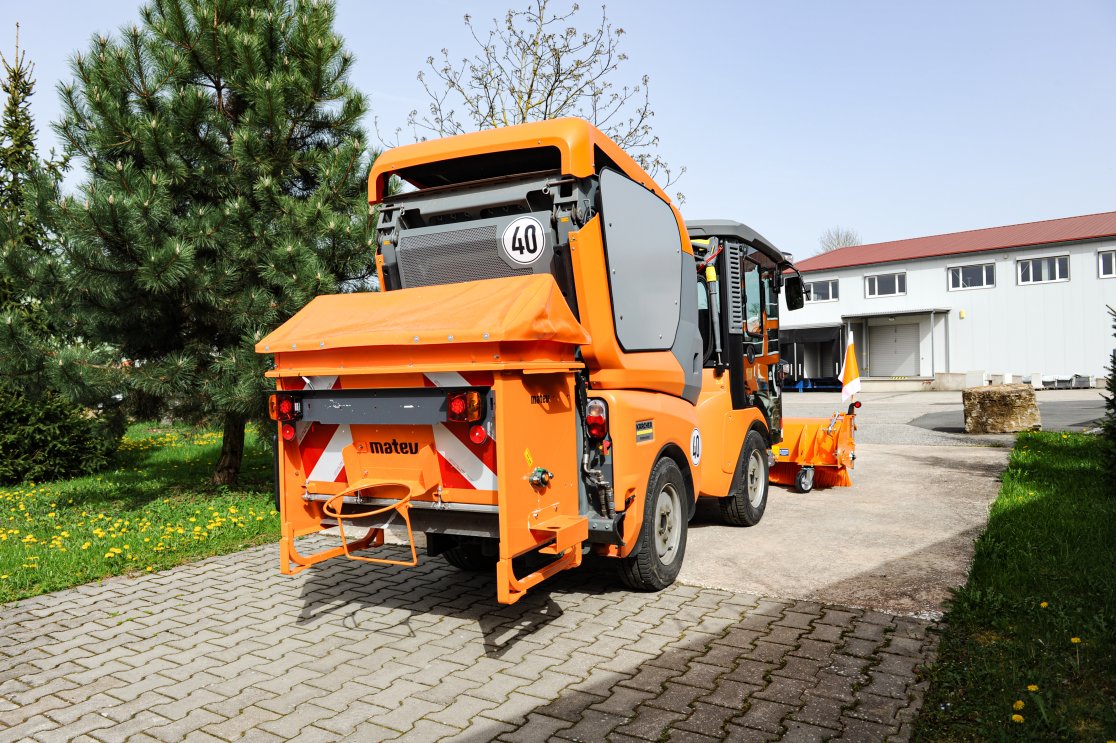 Eine orangefarbene Kommunalmaschine Kärcher MC 130, ausgestattet mit einem matev SPR-BX Kastenstreuer, steht auf einem gepflasterten Hof. Im Hintergrund sind Bäume, eine grüne Wiese und ein Gebäude mit rotem Dach zu sehen.