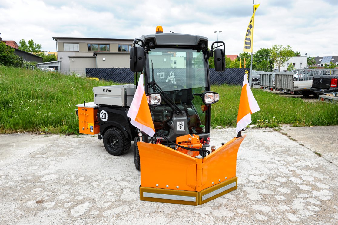 Eine orangefarbene Kommunalmaschine Kärcher MIC 26 mit einem matev SRM-VP Schneepflug und einem Kastenstreuer auf der Ladefläche. Die Maschine ist auf einem betonierten Platz geparkt, im Hintergrund sind eine grüne Wiese, Gebäude und Anhänger zu sehen.