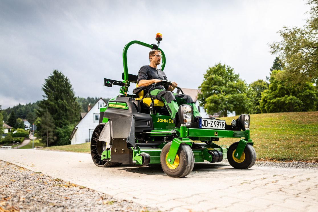 Ein Mann fährt auf einem John Deere Z997R Aufsitzmäher mit StVZO-Kit von matev. Das Fahrzeug ist auf einem gepflasterten Weg in einer grünen, ländlichen Umgebung zu sehen. Im Hintergrund stehen Häuser, Bäume und Hügel.