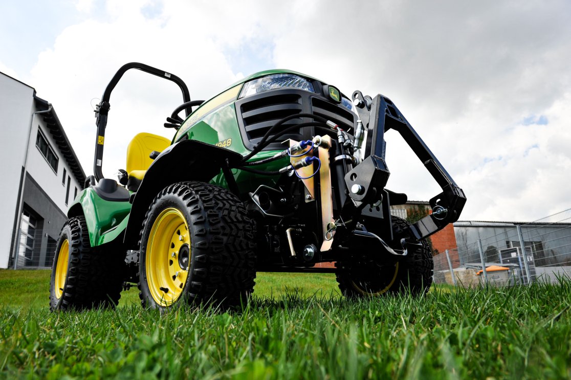 Nahaufnahme eines John Deere X948 Traktors mit montiertem Frontkraftheber. Das Fahrzeug ist grün mit gelben Akzenten, gelben Felgen und robusten schwarzen Reifen. Die Perspektive zeigt das Fahrzeug aus einem niedrigen Winkel, während es auf einer grünen Wiese steht. Im Hintergrund sind Gebäude und ein bewölkter Himmel sichtbar.