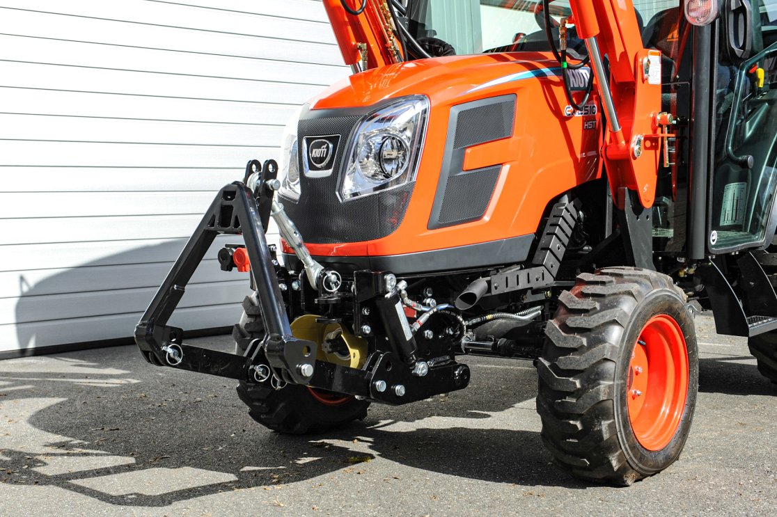Nahaufnahme eines KIOTI CX2510 Traktors mit angebautem matev Frontkraftheber. Der Traktor in leuchtendem Orange steht auf einem asphaltierten Boden vor einer weißen Garage. Die kräftigen Vorderreifen und der Frontkraftheber sind gut sichtbar.