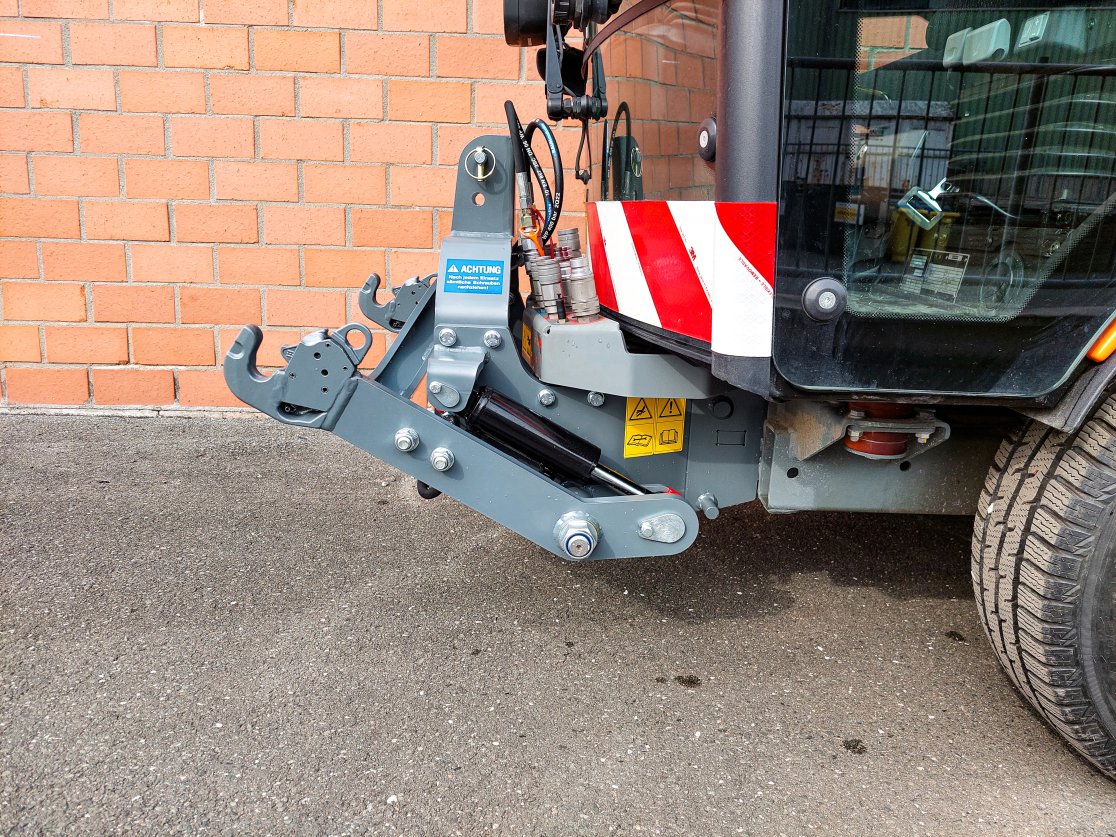 Detailaufnahme einer matev FPS-Frontaushebung an einer Kärcher MC 130 Kommunalmaschine. Die graue hydraulische Einheit ist an der Front des Fahrzeugs angebracht, vor einer roten Backsteinwand.